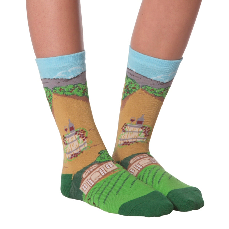 K Bell American Made Womens Winery Sock starts on a Green background and has Dark Green Toes and Heels. The Vineyards are in Rows up and down from the main house. The mountains rise up to a Blue Sky. This sock is on a Model.
