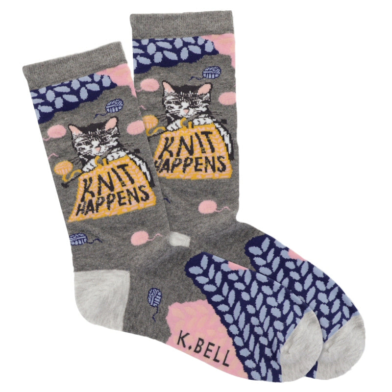 K Bell Knit Happens Sock starts on a Charcoal Heather background. The Heels and Toes are Silver Heather. Navy Blue and Baby Blue, and Coral and Coral Pink are colors of Balls of Yarn around the Sock. Under the Cuff is a Cute Knitter, a Kitten. Charcoal Grey and White with Black. The Kitten has Coral Pink Ears and Nose. is holding knitting needles in her paws. She has Knitted a Blanketl and with Gold Lurex reading "Kint Happens".