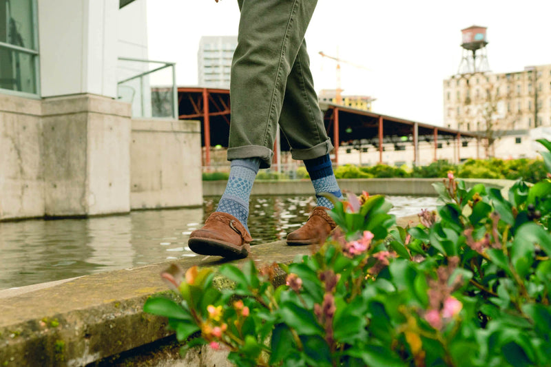 Model wearing green pants, Solmate Horizon Socks with brown shoes standing on a concrete ledge with flowers and water. 