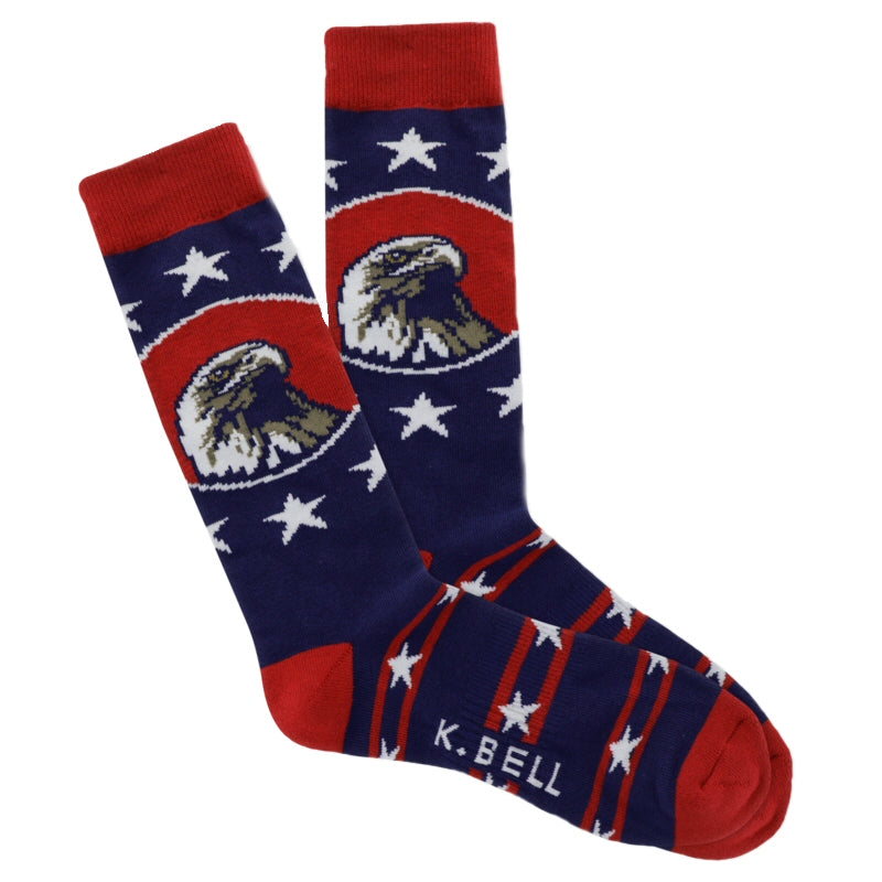 K Bell American Made Mens Bald Eagle Sock starts on a Navy background with Red Cuffs, Heels and Toes. The American Bald Eagle is in the center of a Red Oval in a Portrait. Around Him are Five Point Large White Stars in a Field of Blue.  On the Foot are Blue Bars of smaller Five Point White Stars all around with Red small Stripes making each Bar.