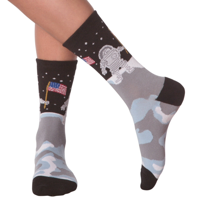 K Bell American Made Womens Man On The Moon Sock is started with Black background on the Toes, Heels and Cuffs. Under the Cuff above the Horizon of the Moon is the Blackness and White Stars. The Astronaut is holding the American Flag ready to plant it in the Lunar soil. The soil is Medium Grey, Light Blue and White. This Sock is on a Model.