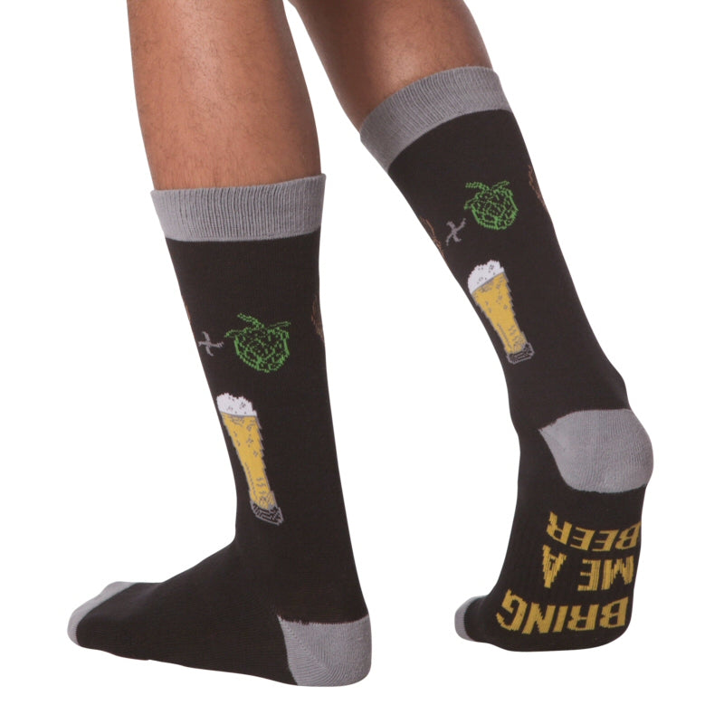 K Bell American Made Mens Beer Sock is shown on a Model. It starts on a Black background with Charcoal Grey Cuffs Heels and Toes. Under the Foot reads, "Bring Me A Beer" in Bold Golden Text. Below the Cuff are Brown Barley then a Plus Sign and Green Hops. Below is the Equals of a Glass holding a Beer, Golden with a Head of White Foam. This is a Large Sock. 
