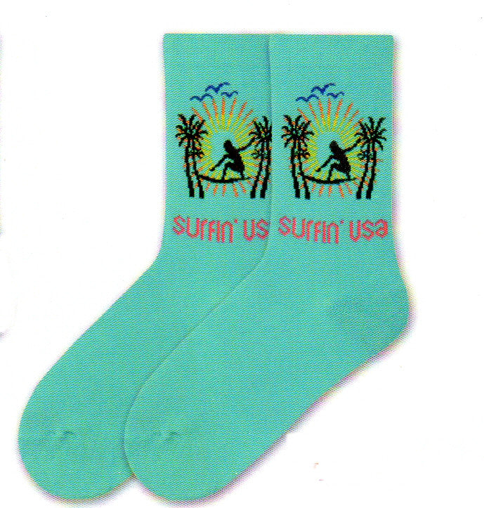 K Bell American Made Womens Surfin' USA is on a background called Jewel. A Silhouette of a Girl in Black is on a Surf Board. With Bright Yellow and Orange streams of light. Blue Silhouette of birds. To finish off the scene are Black Palm Tree Silhouettes. 