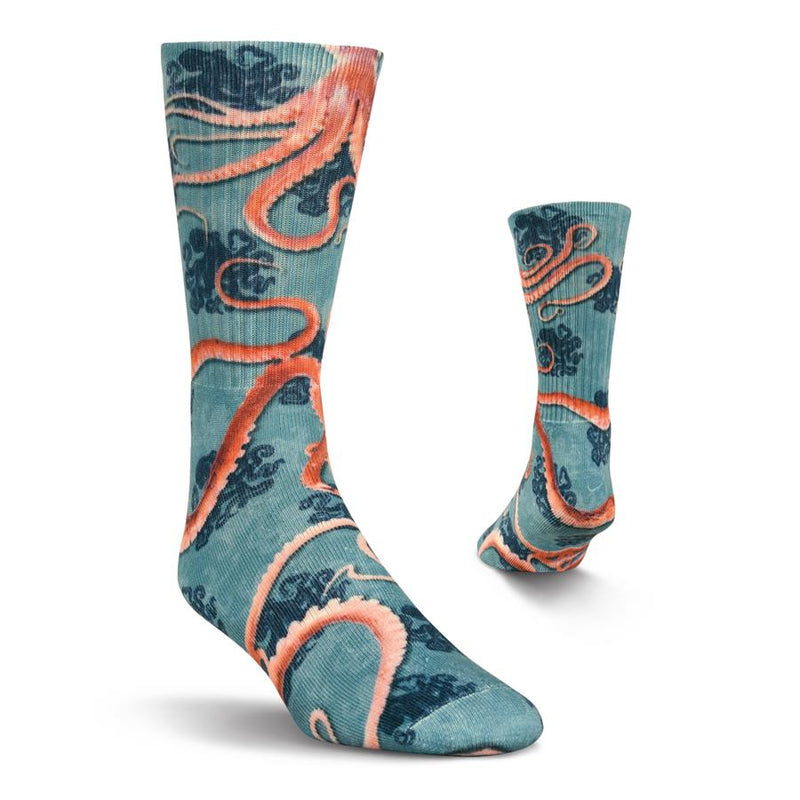 Kurb Mens Octopi Crew Sock starts on a Blue background. Light Blue and Navy Silhouettes are all around the Sock. Then 8 arms crawl over the Sock in Reddish Brown and Coral Colors. At the Cuff are Two Octopi.