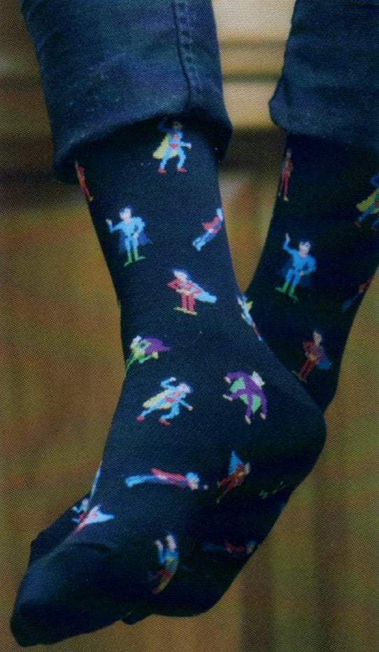 K Bell Mens Super Heroes Sock starts on a Black background.  From the Cuffs to the Toes are Super Heroes in different colored outfits and capes.  Some are flying, standing and running. Colors are Bright and pop off the Black in Red, Blue, Yellow. Green and Purple. This Sock is shown on a Model.