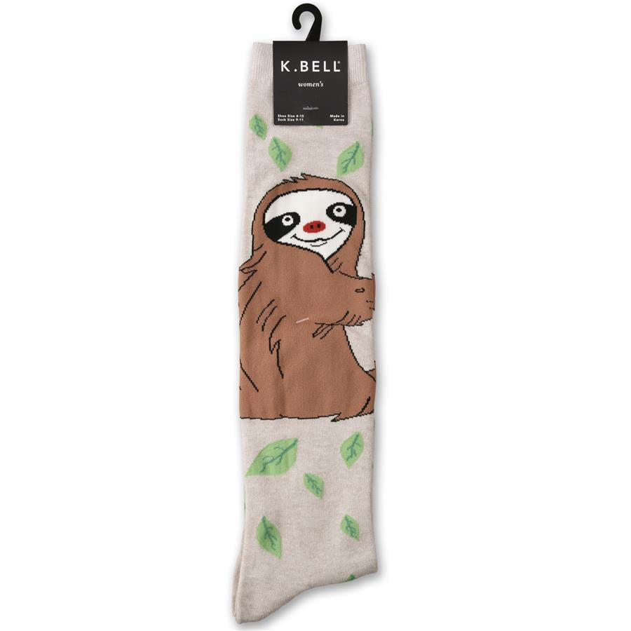 K Bell Silly Sloth Knee High Sock is on a background of Oatmeal Heather. Green Leaves are all around. A Three Claw Sloth sits upright in Brown, Sandy Brown with a Black Eye Mask and Mouth. The Nose is Turkey Red.