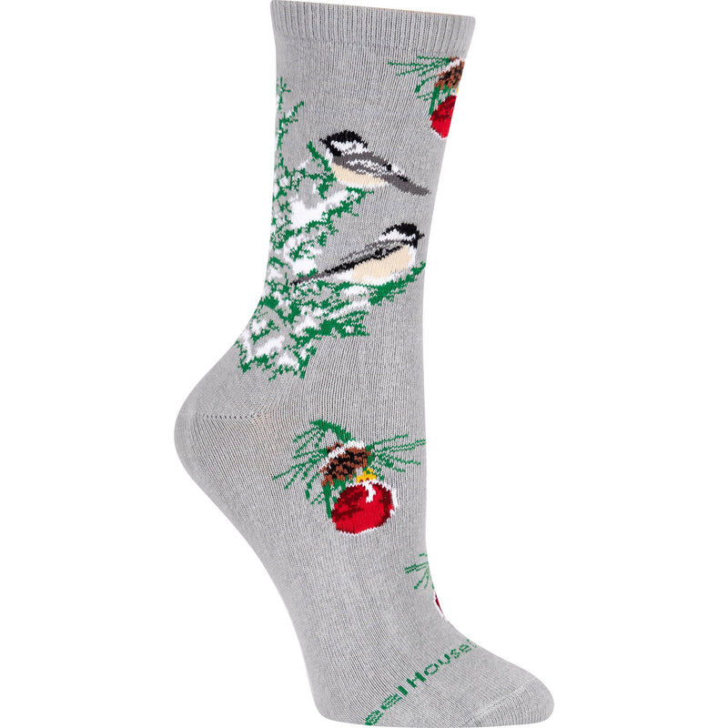 Wheel House Designs Chickadee Christmas Tree Sock is on a Grey background with branches of a snow covered Christmas Tree. On the Tree are Chickadees perched. They are Black White Dark Grey and Vanillia.  Above and Below are Sprigs of the Tree with Pine Cones and a Red Ornament.